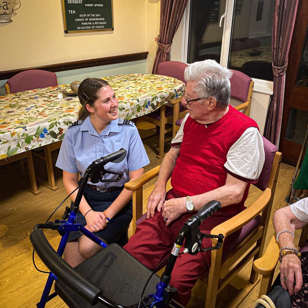 Smiling Female air cadet sits beside sitting elderly care home resident as they chat.